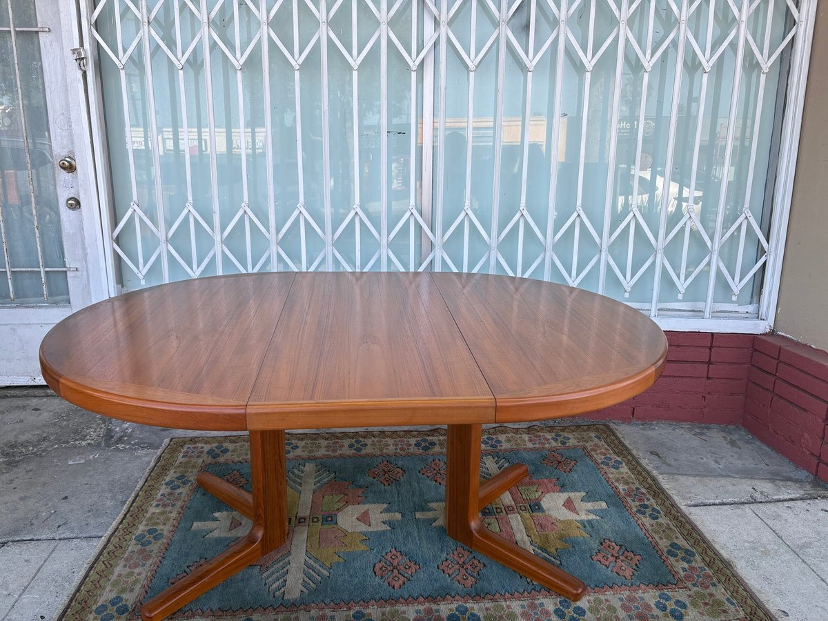 Oval wooden dining table with pedestal base on a patterned rug, positioned in front of a metal security door
