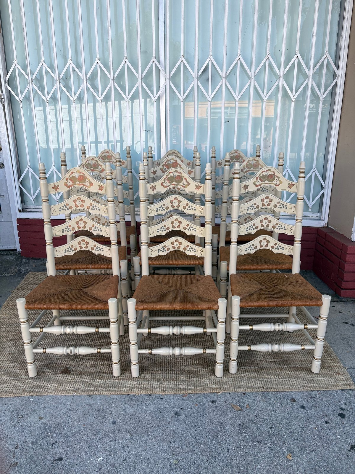 Six white wooden chairs with rush seats arranged in two rows on a woven mat in front of a window with white accordion blinds.