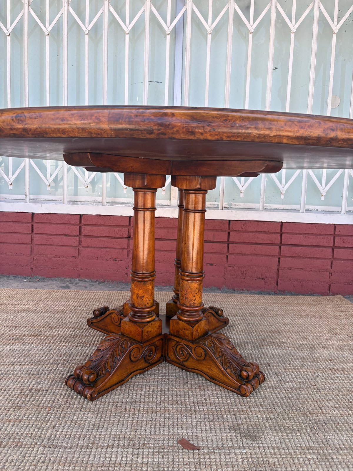 Antique wooden pedestal table with ornately carved legs and cross-base feet, displayed outdoors against corrugated metal and brick wall background