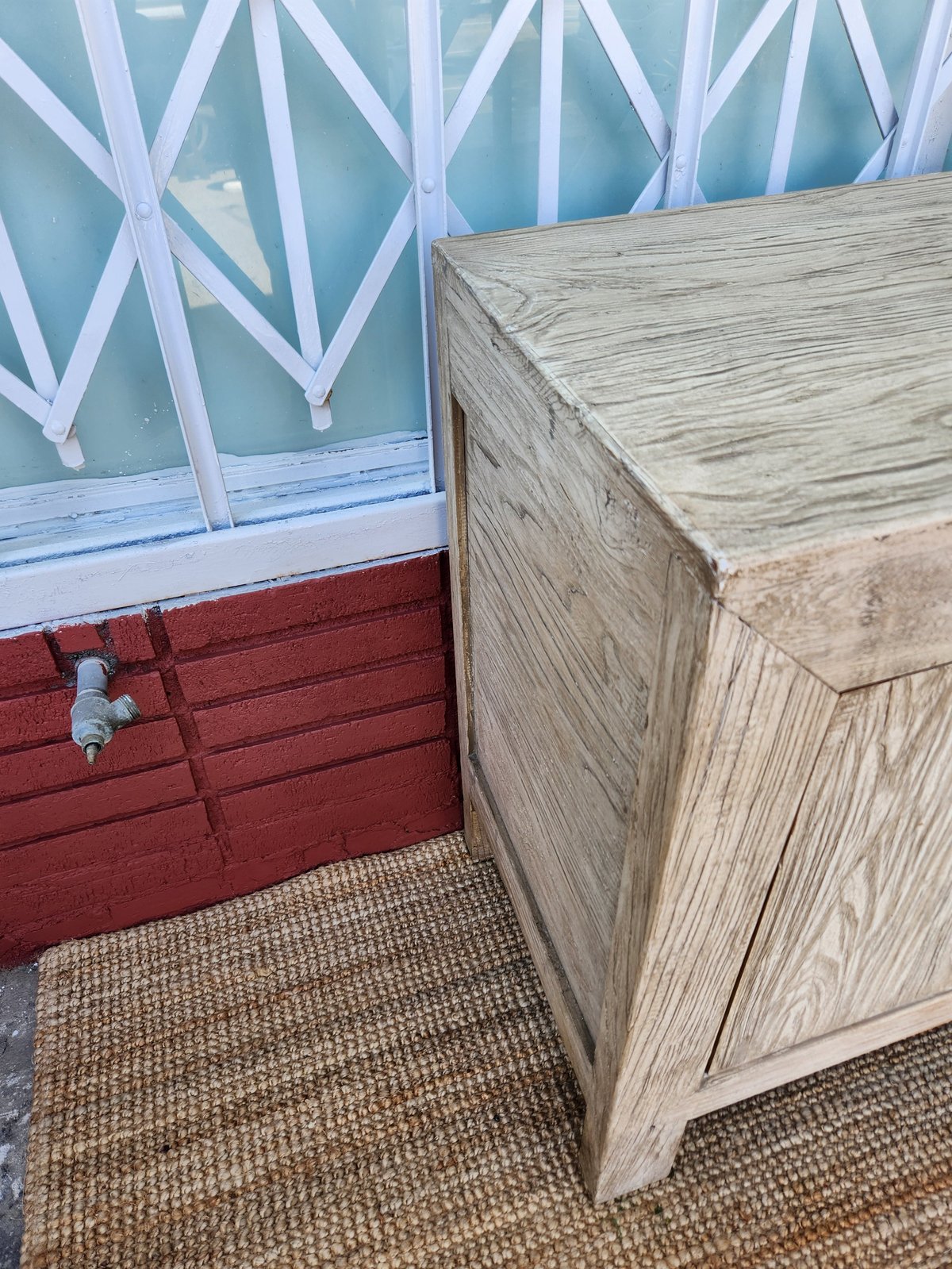 Interior of a beach hut with turquoise window, red-painted lower wall, white striped curtains, wooden bench, and sisal flooring