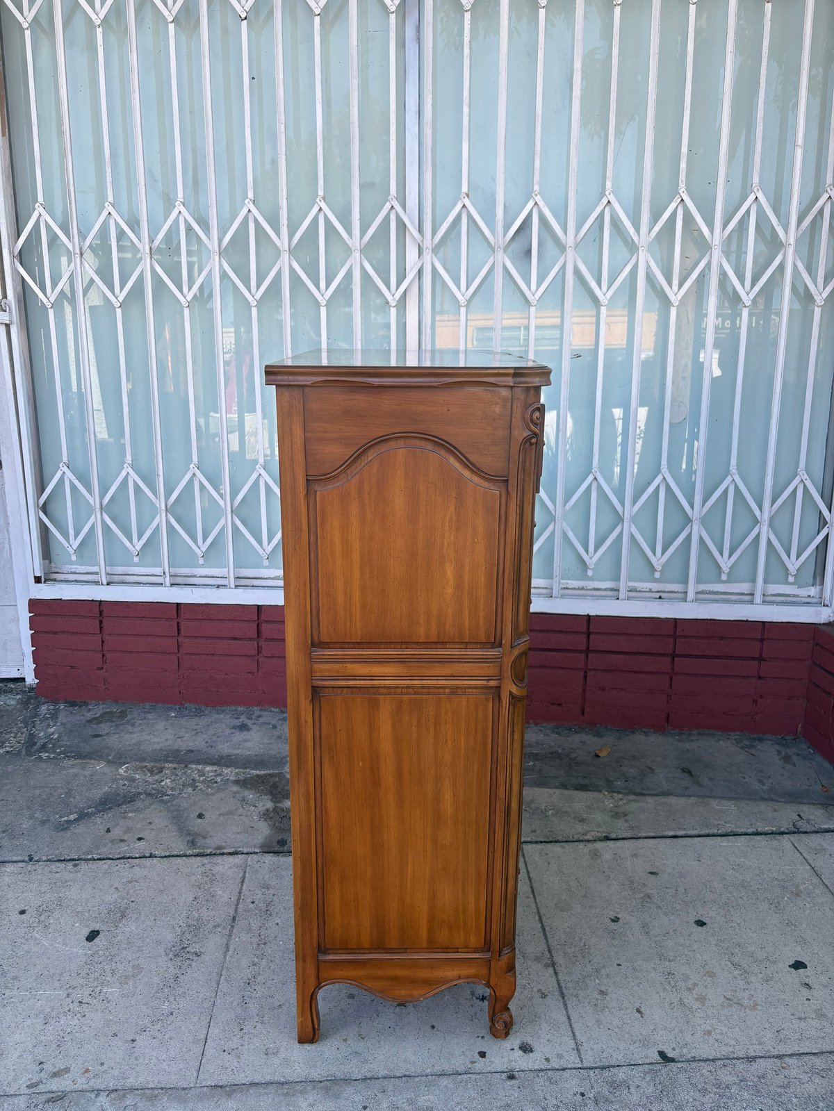 Wooden cabinet with two doors on a sidewalk in front of a metal accordion gate with red base