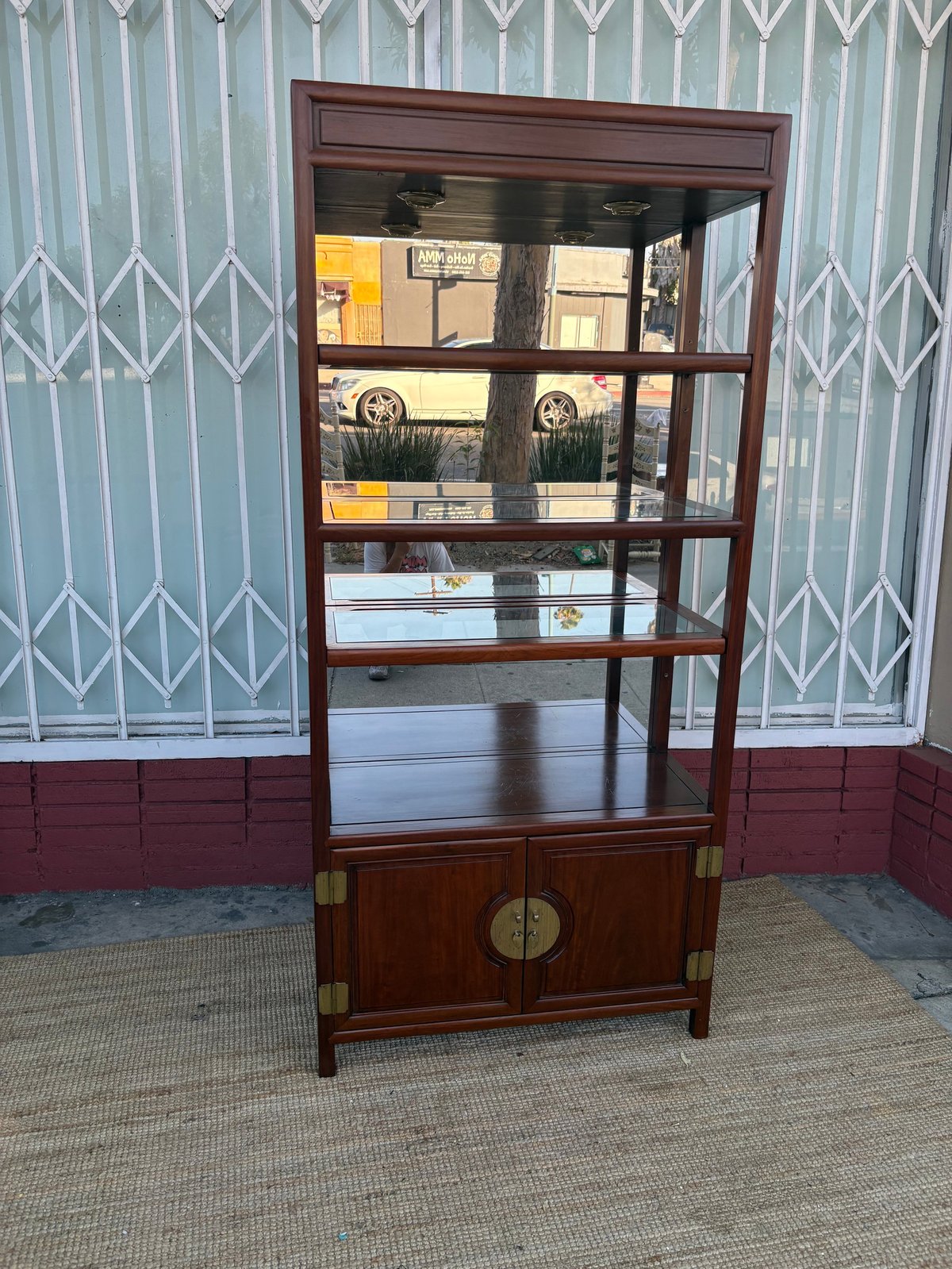 Asian-inspired wooden display cabinet with glass shelves and lower storage doors against an ornate metal gate backdrop.