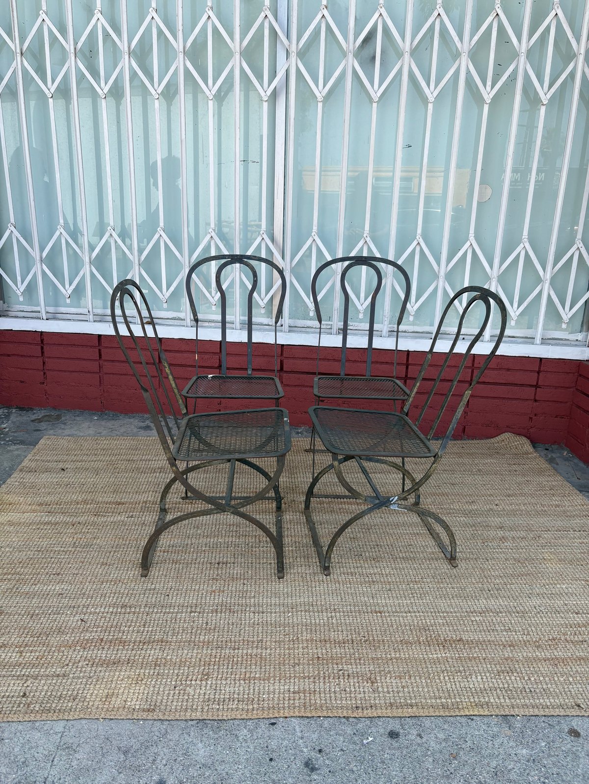 Four metal chairs arranged around a table on a beige outdoor rug against a storefront with a metal grill gate and red wall