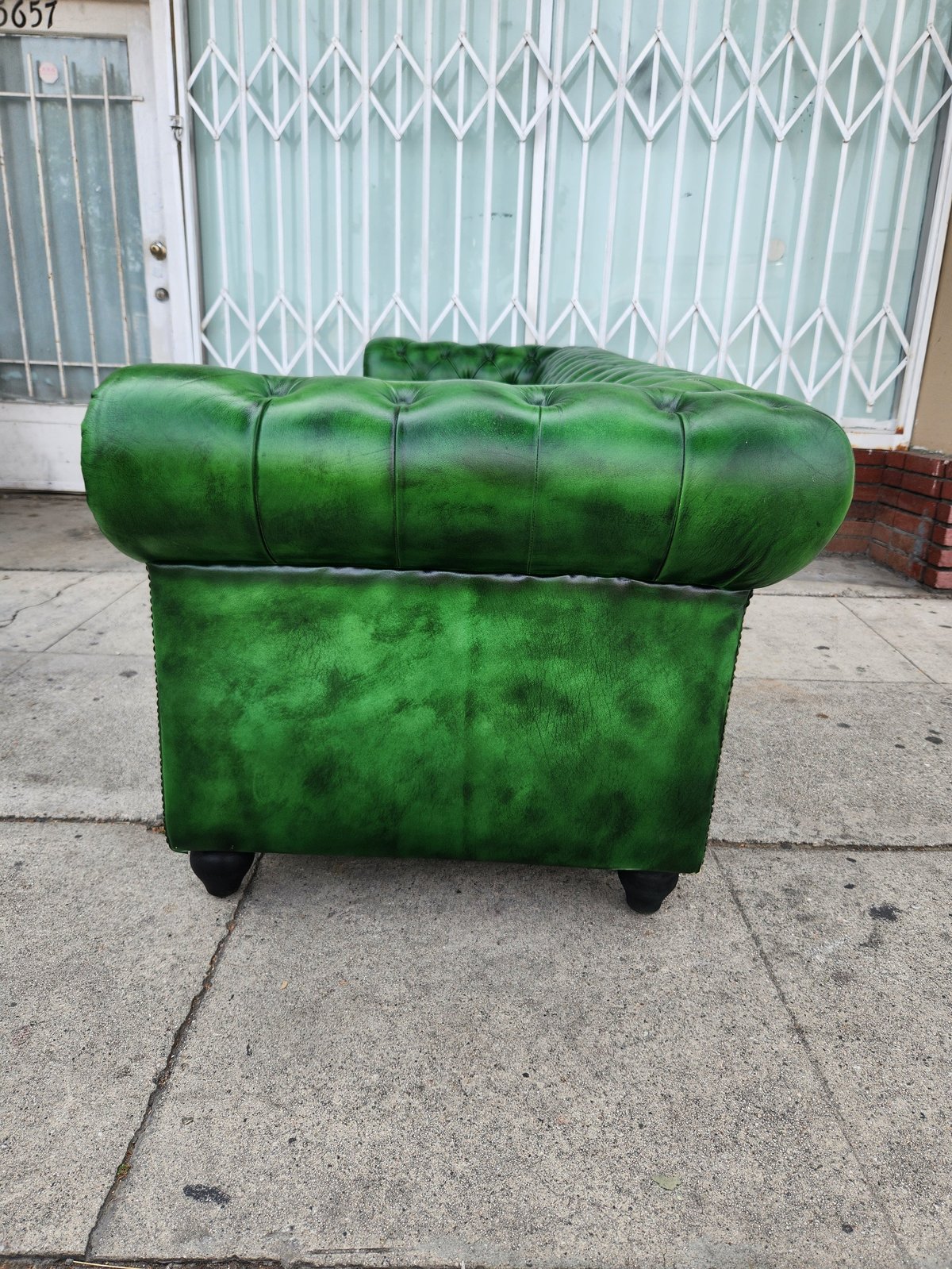 Green velvet tufted armchair sitting on a driveway in front of a metal gate and white storefront