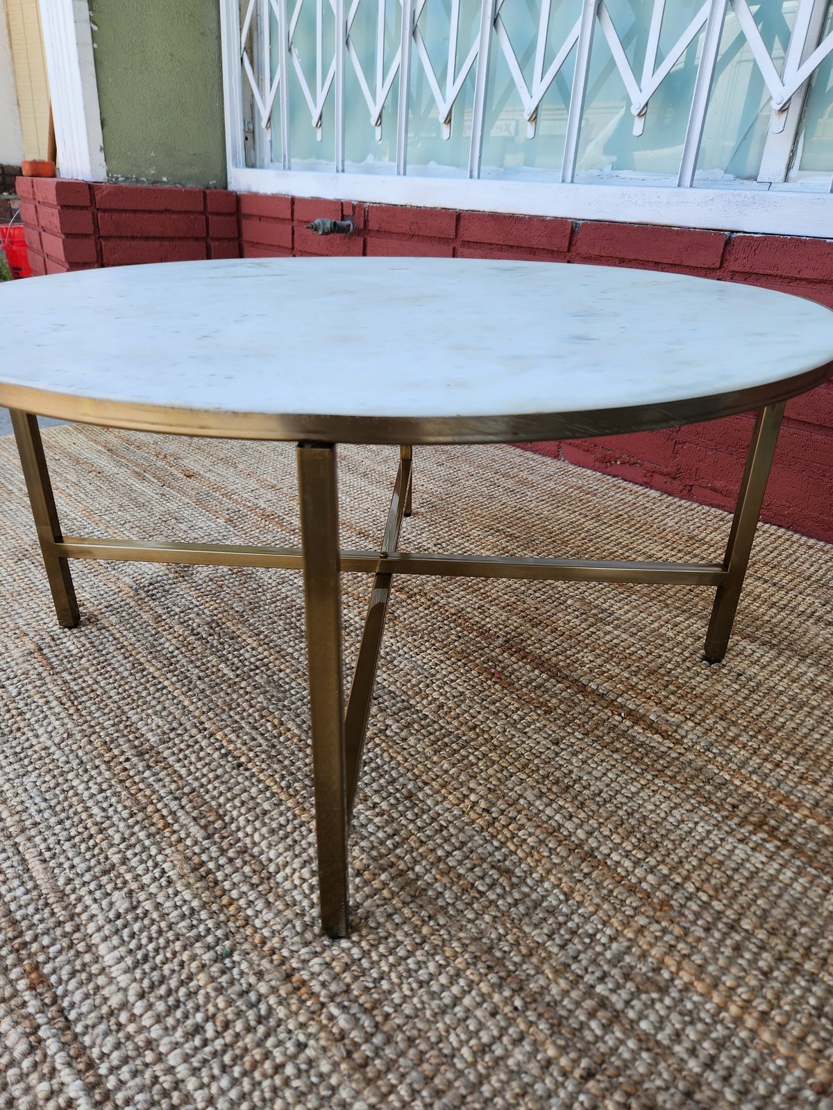 Oval marble-top table with bronze metal legs on beige carpet in a room with red brick wall and window