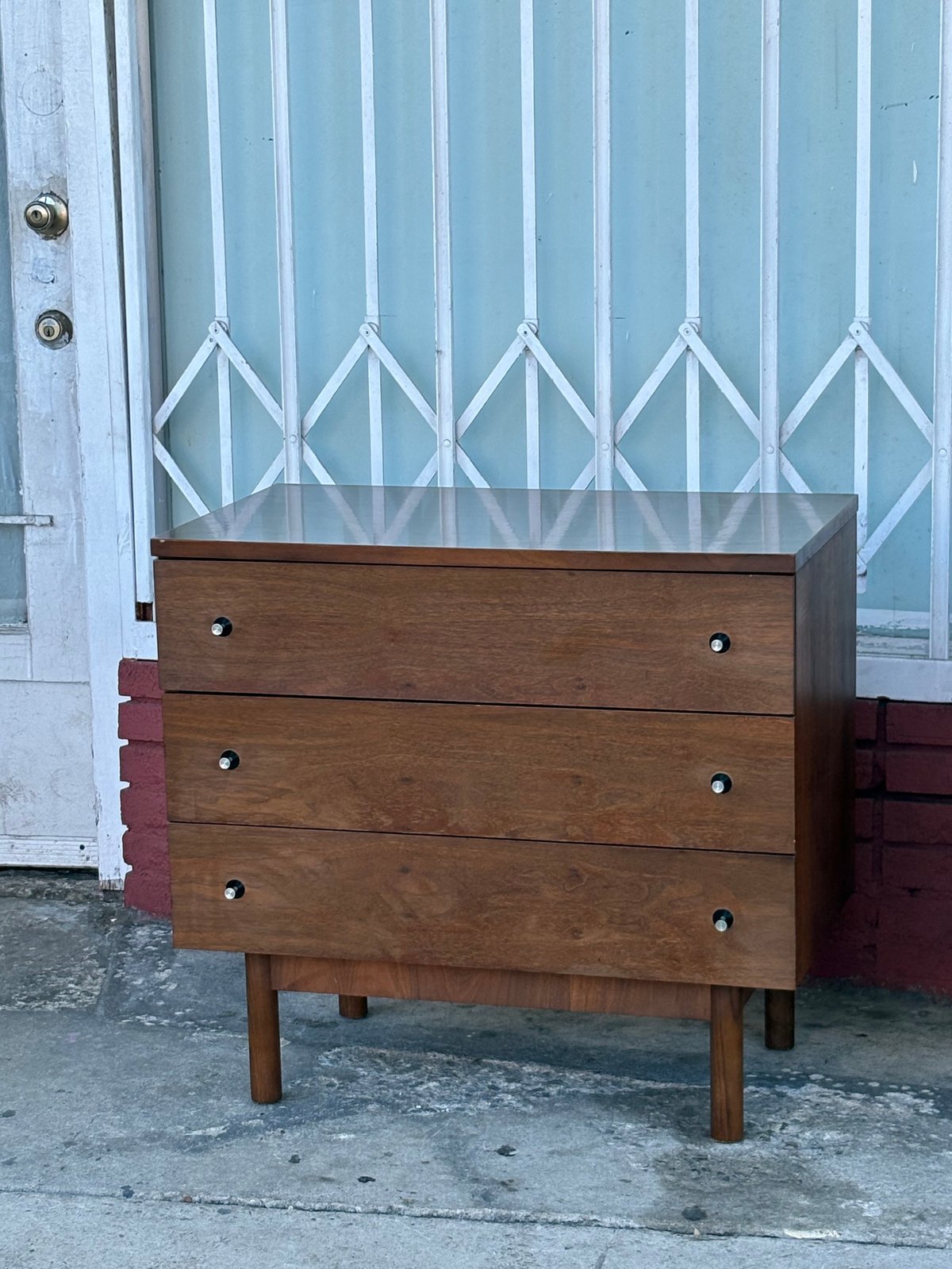 Mid-century wooden dresser with three drawers positioned against a light blue corrugated metal wall