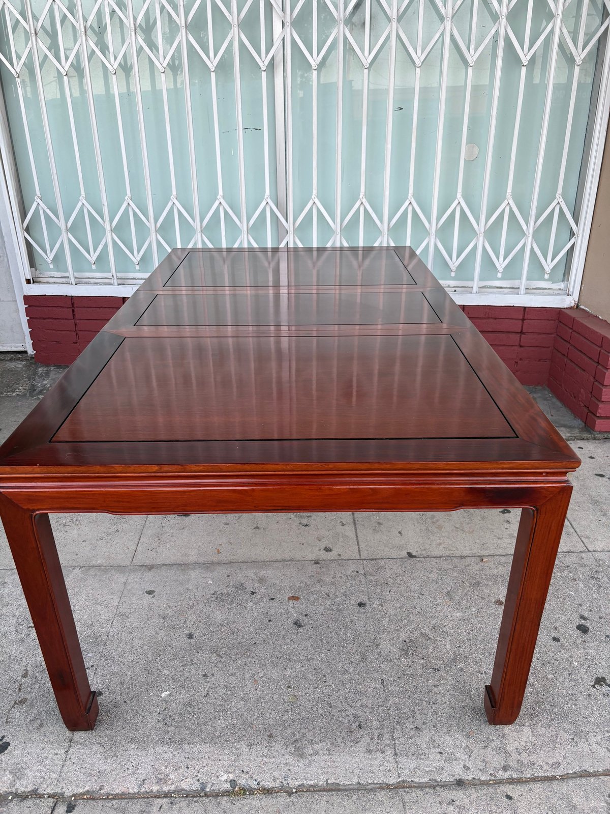 Wooden dining table with glass top surface in front of white metal gate and red brick wall