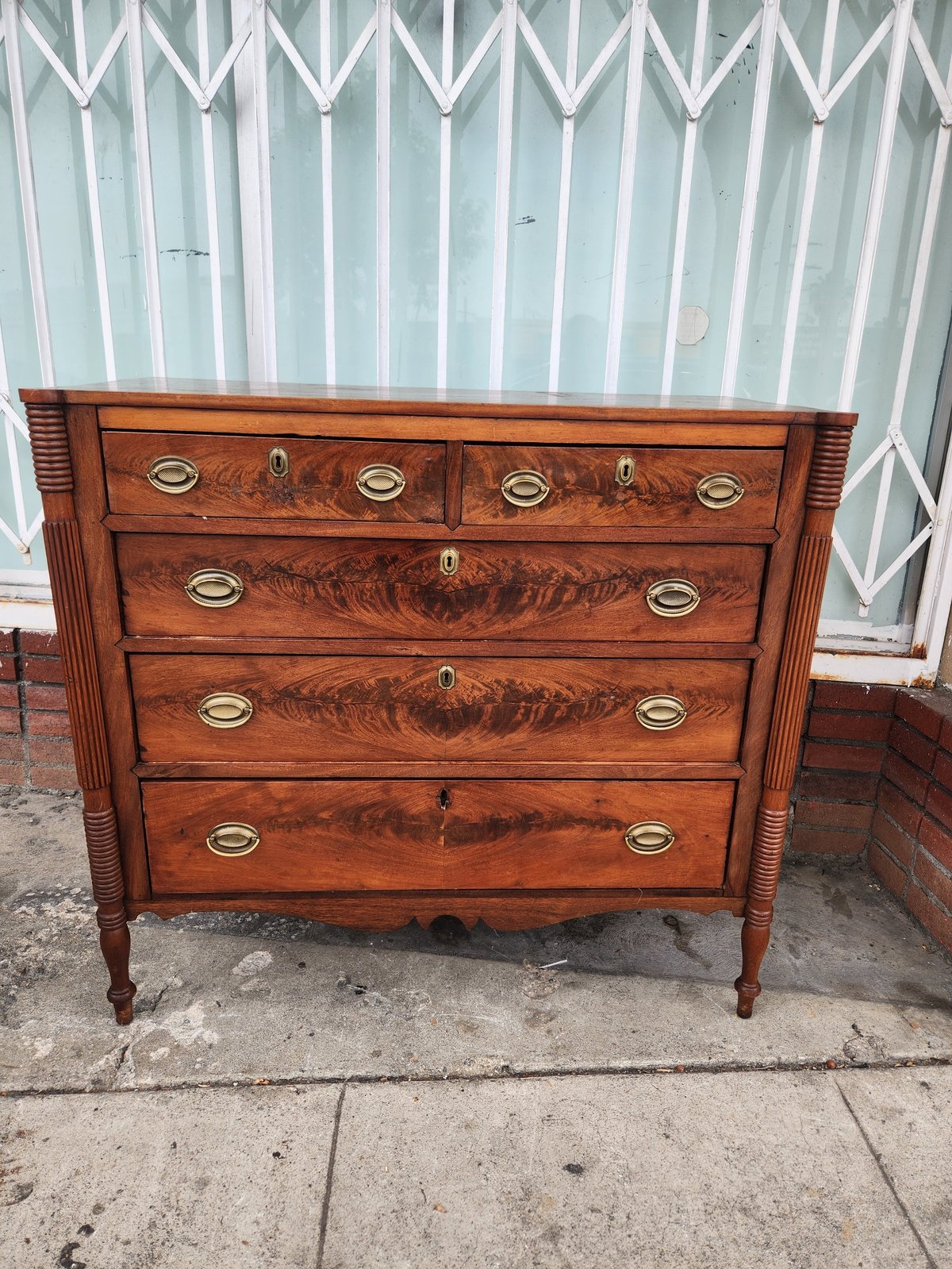 Antique wooden dresser with five drawers and brass handles on a concrete surface against a corrugated metal wall