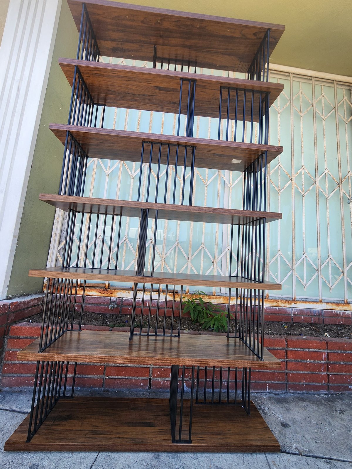 Tall wooden shelving unit with black metal cage-like frames on each level, displayed against a yellow wall with decorative patterns