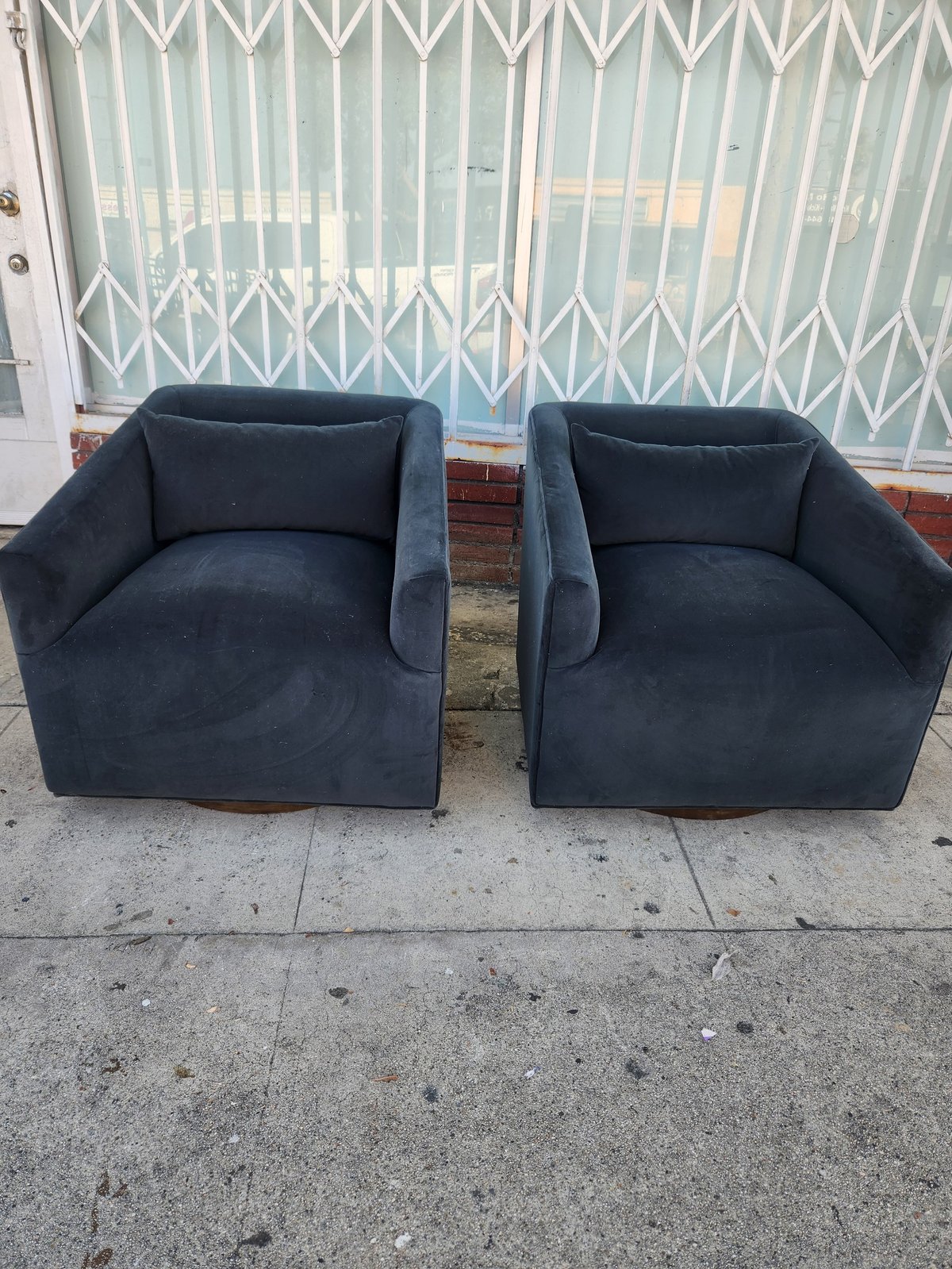 Two dark blue upholstered armchairs positioned on concrete sidewalk in front of white metal gate and brick building