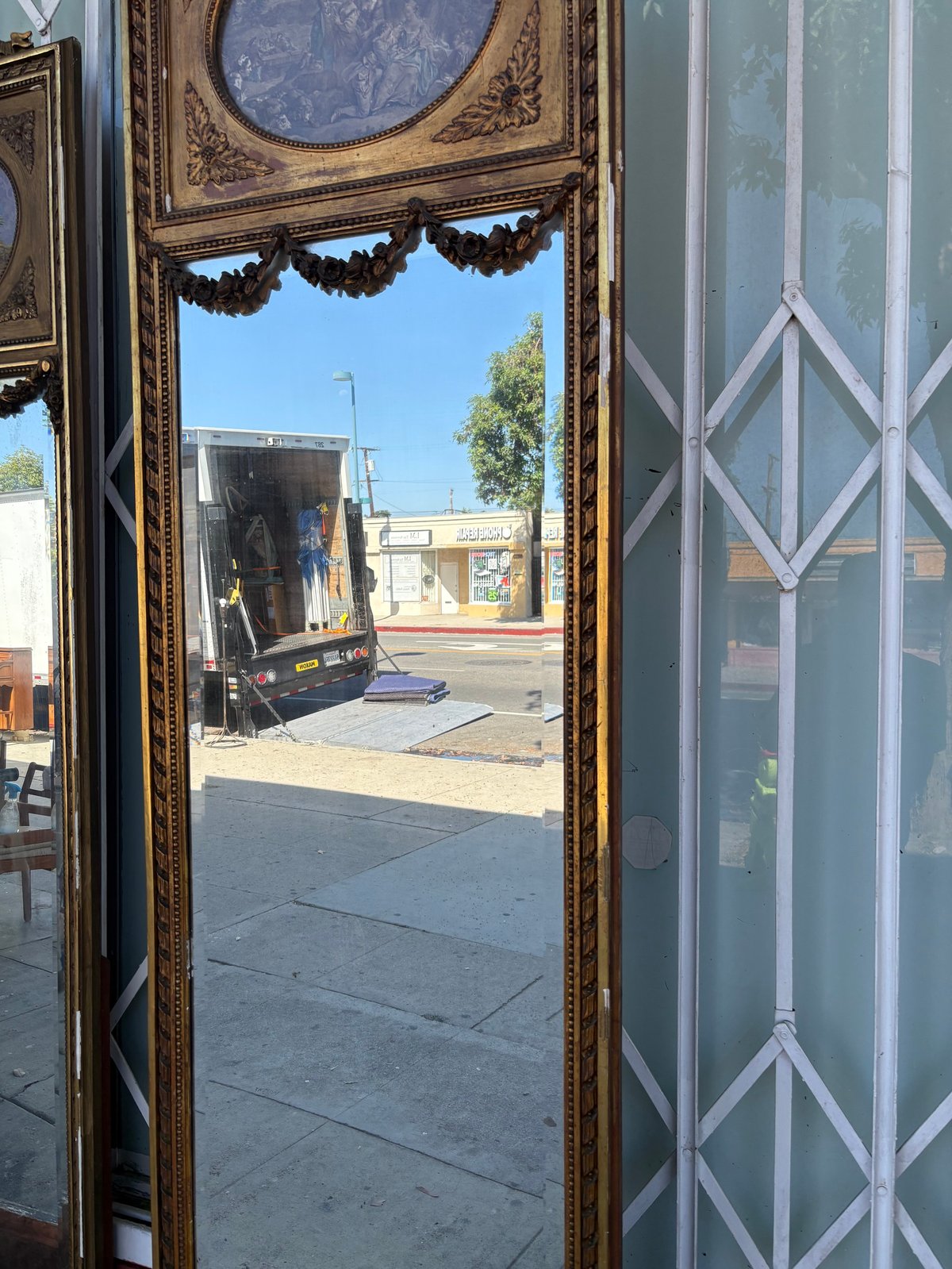 Ornate gold-framed mirror reflecting a sunny street storefront with vehicles and buildings visible in the reflection
