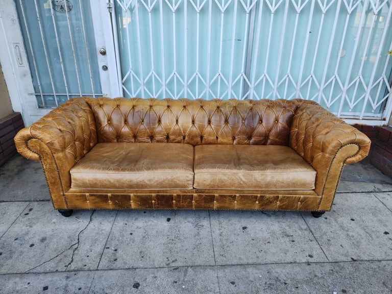 Tan leather Chesterfield sofa with button tufting displayed on a sidewalk against a blue metal gate