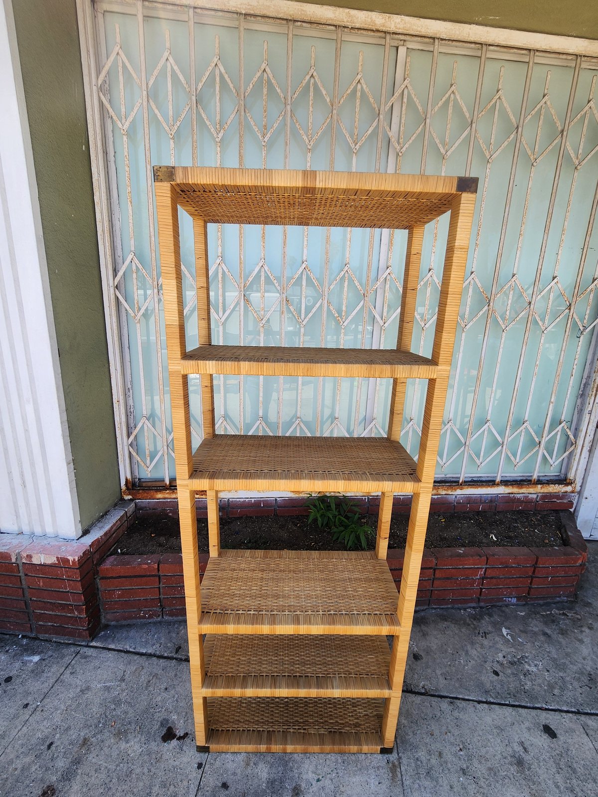 Wooden bookshelf with woven rattan shelves against a decorative glass door and brick planter