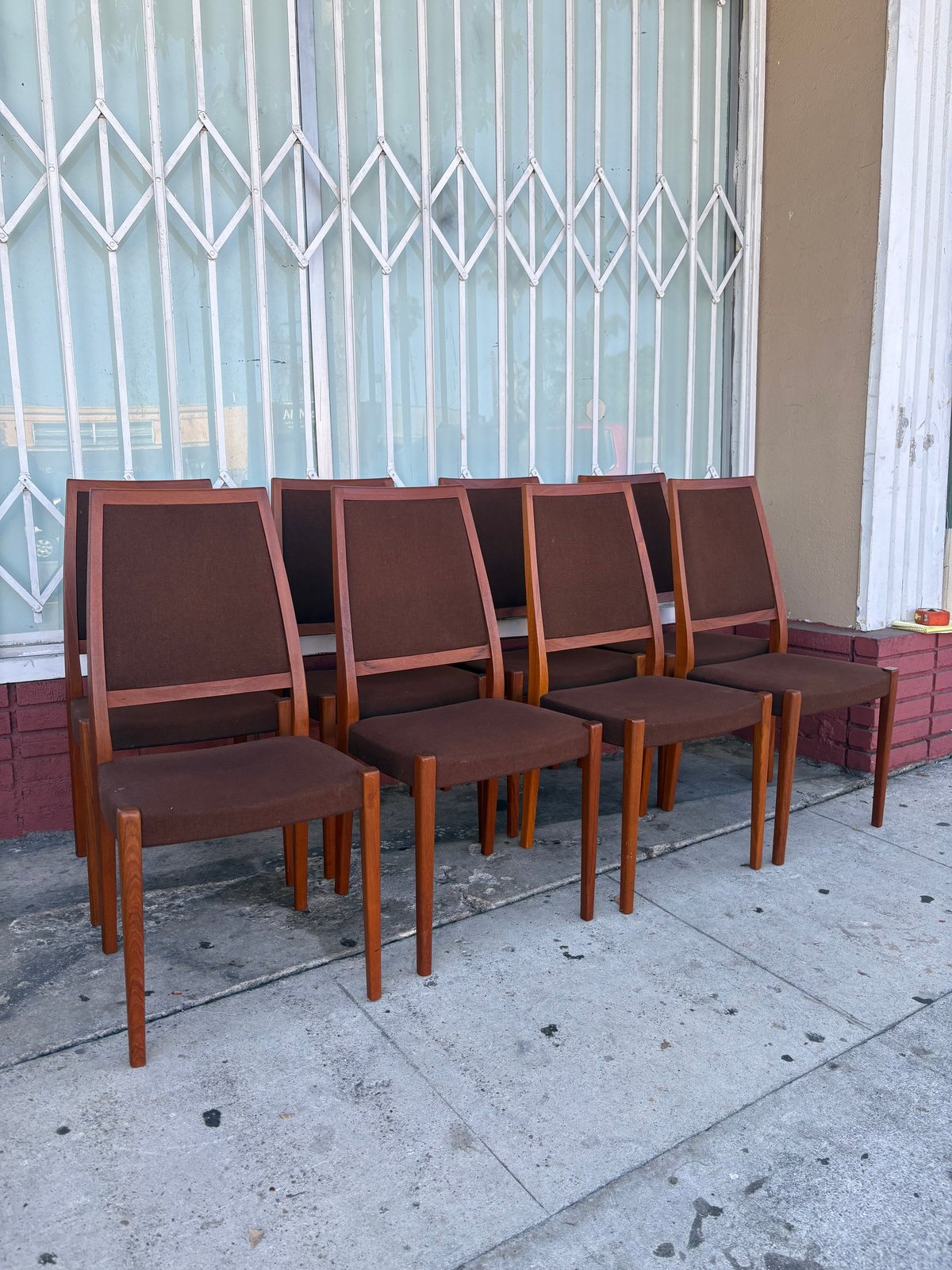 Four mid-century modern dining chairs with brown upholstery and teak wood frames arranged on a concrete sidewalk