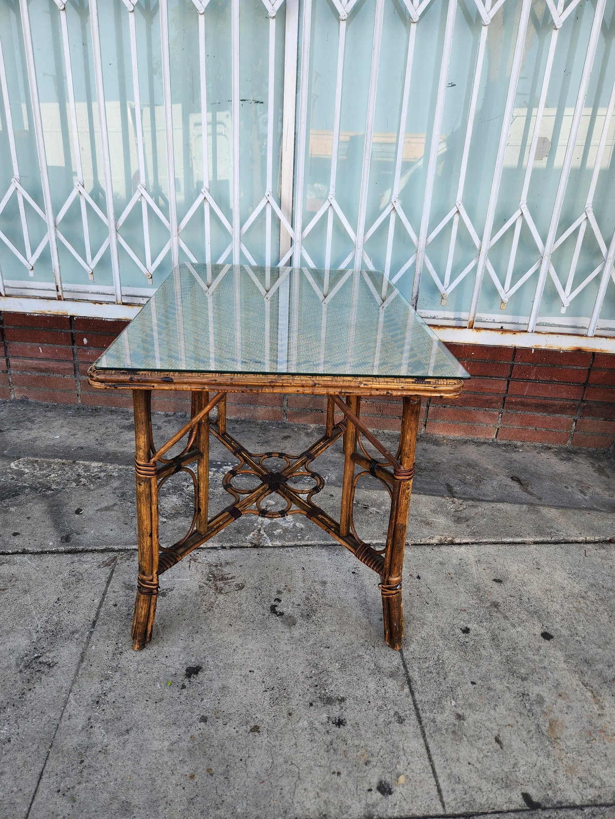Glass-top bamboo table with crossed X-pattern base on concrete pavement in front of white accordion gate