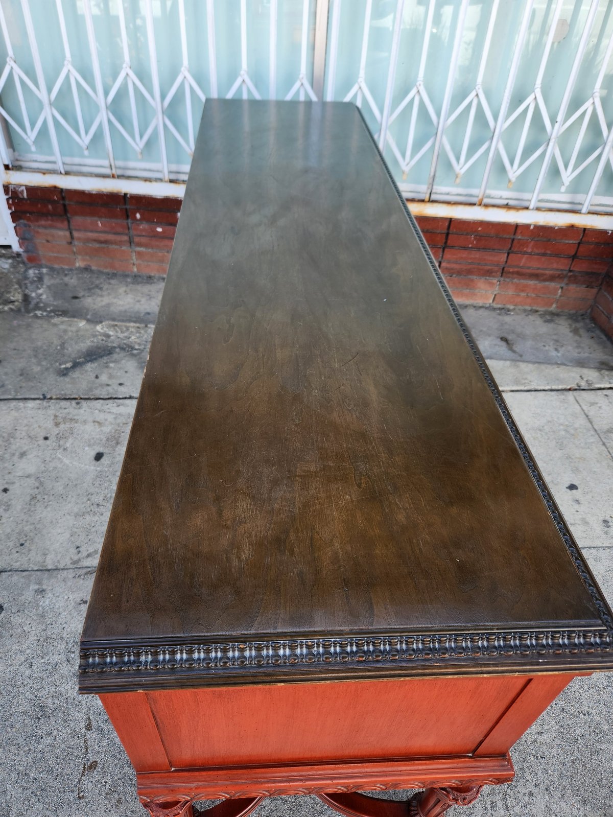 Antique wooden dining table with dark top and red painted base, displayed outdoors against a brick wall and white fence