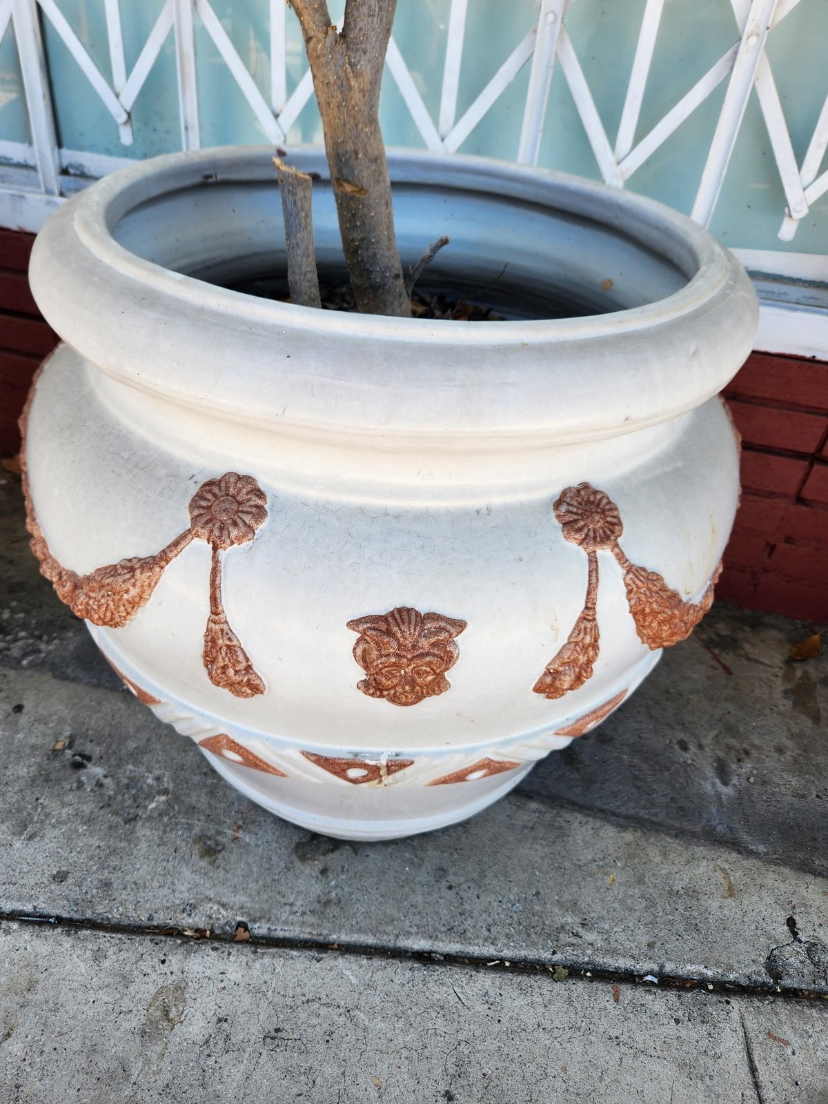 White ceramic planter with brown botanical motifs, containing a small tree, positioned against a decorative turquoise wall