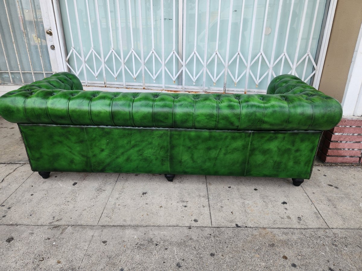 Green tufted Chesterfield sofa positioned on a sidewalk in front of white blinds and a beige wall