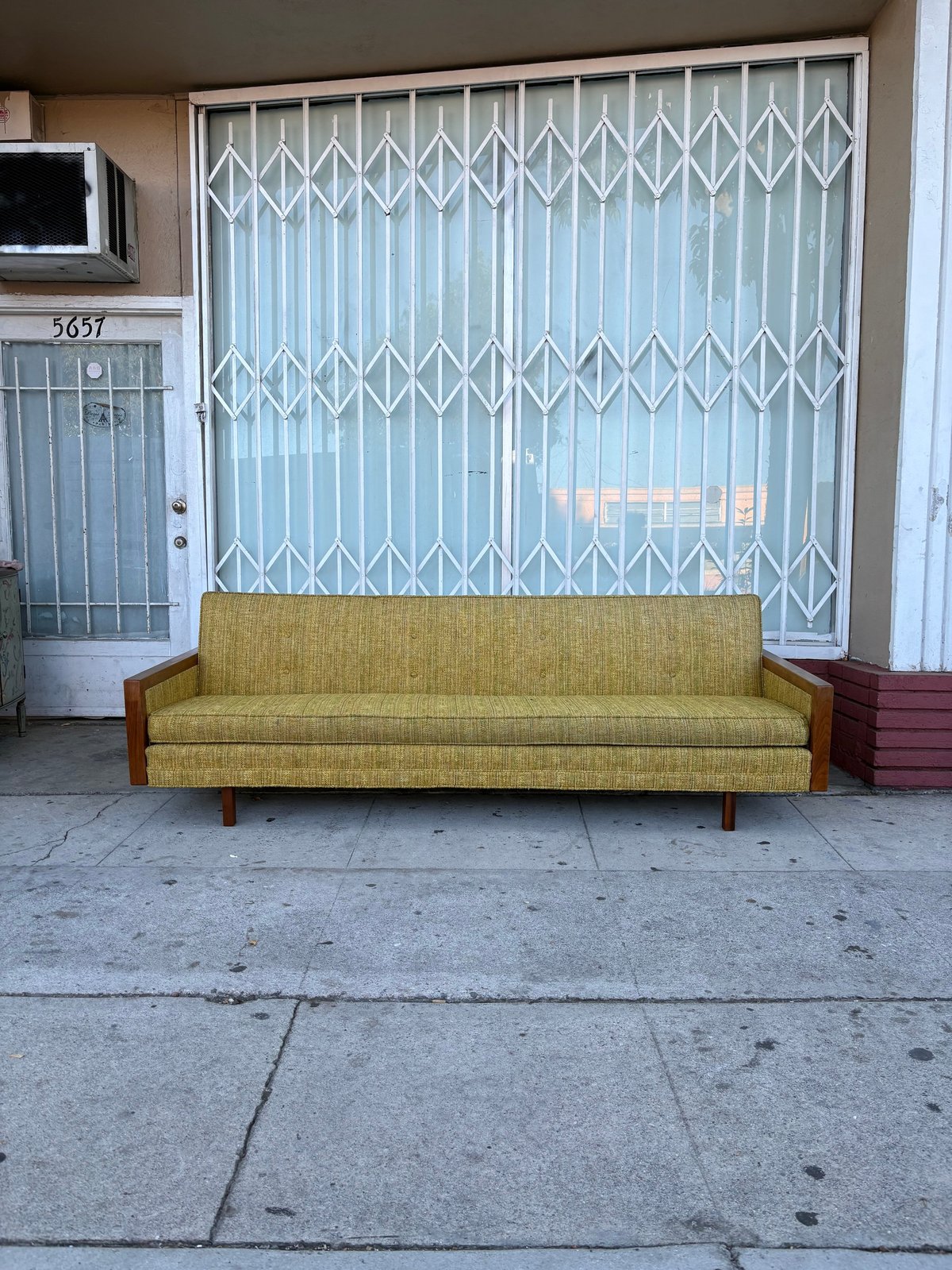Yellow-green mid-century modern sofa on a sidewalk in front of a storefront with metal security gates