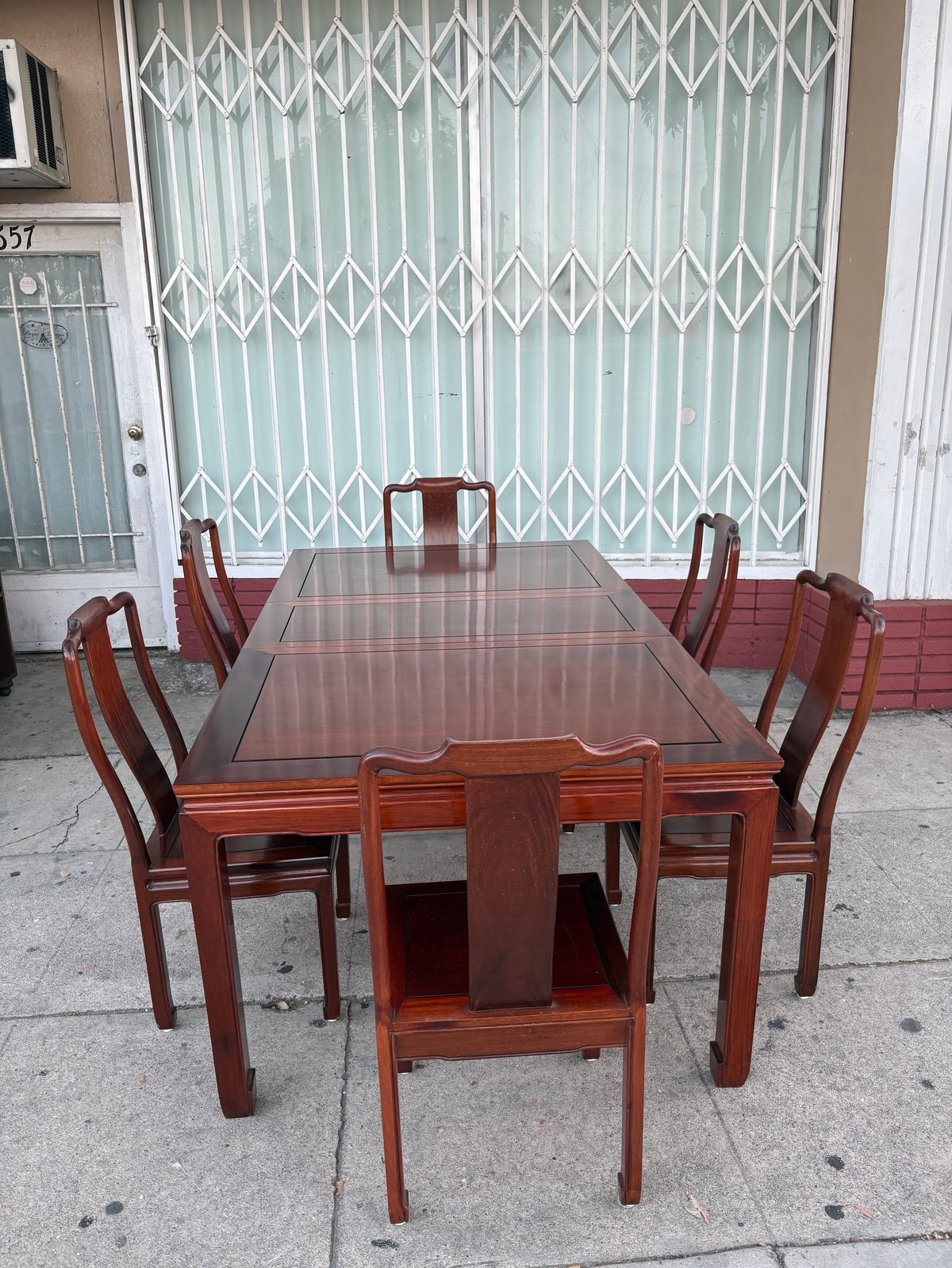 Wooden dining table set with six red chairs positioned on a concrete sidewalk in front of a metal gate.