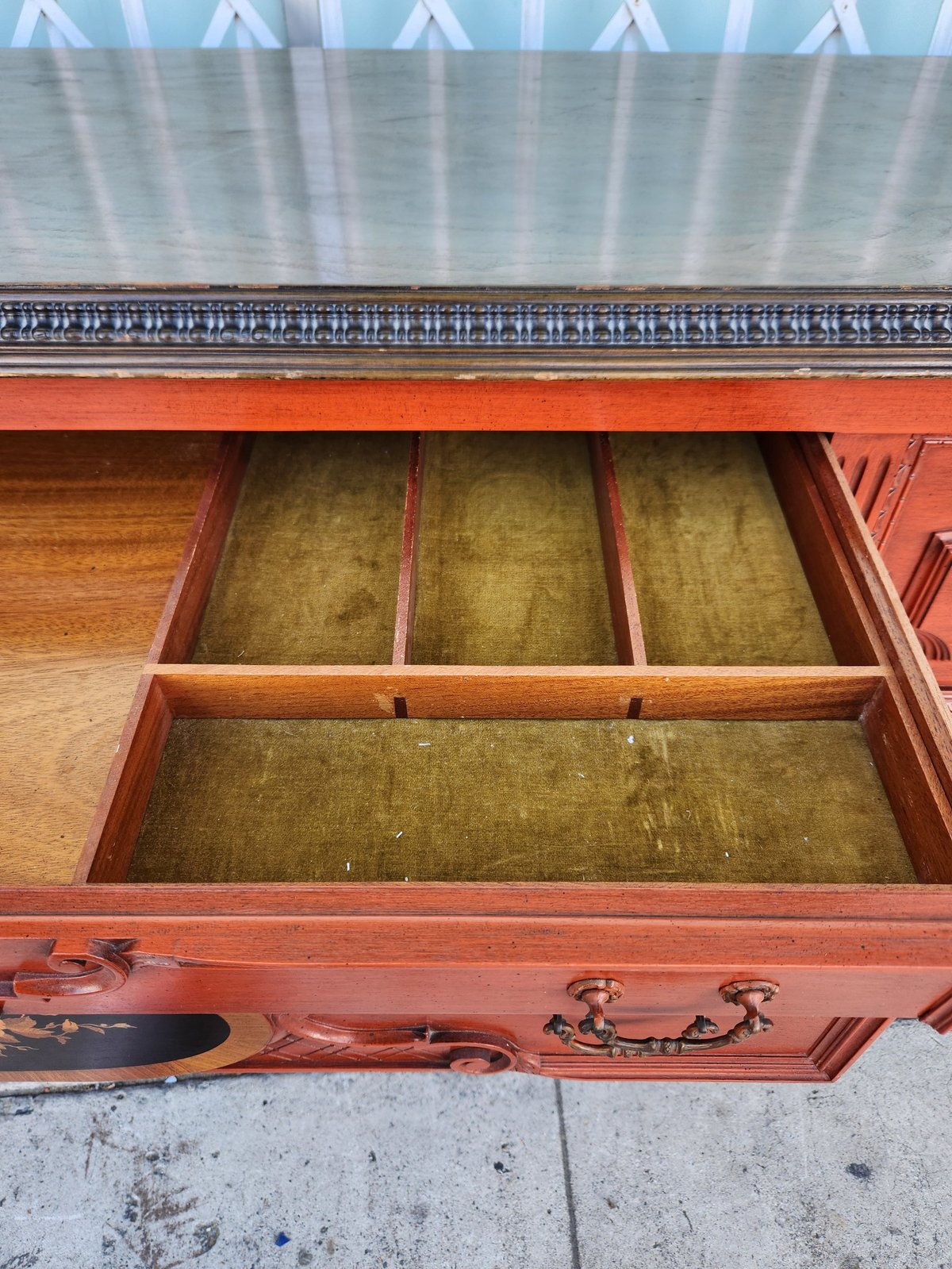 Open red-orange wooden dresser drawer showing interior compartments with brass-colored lining and ornate metal hardware