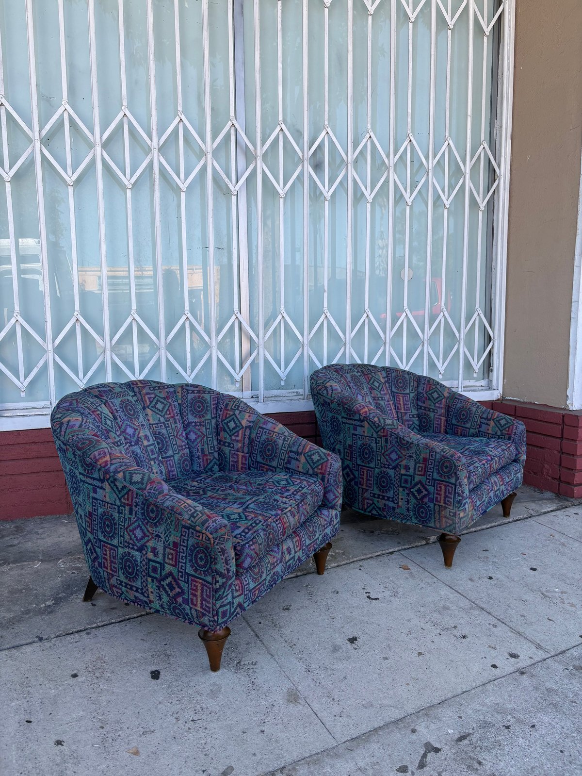 Two patterned upholstered armchairs on a concrete sidewalk in front of a white accordion gate and red brick building