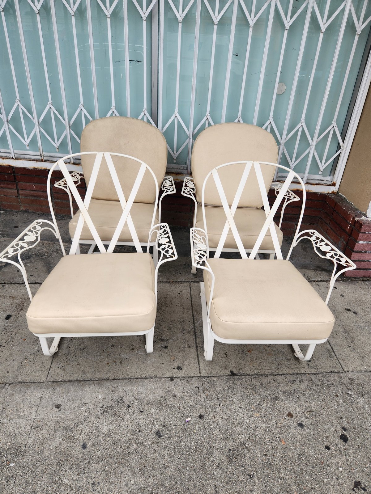 Two white metal outdoor lounge chairs with beige cushions positioned on a concrete sidewalk in front of a teal patterned window shutter