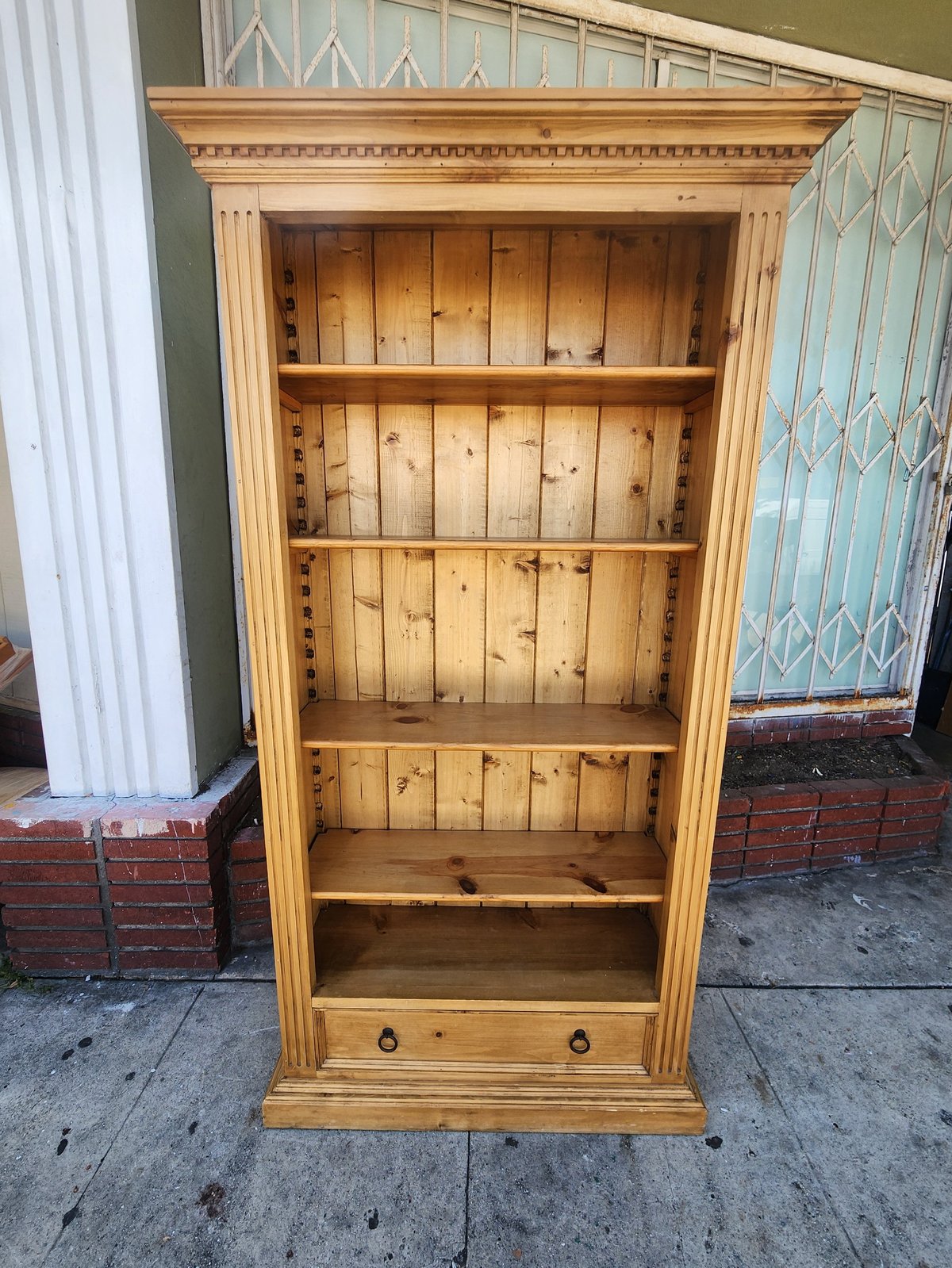 Wooden bookshelf with four shelves, decorative top, and bottom drawer sitting outdoors on concrete ground