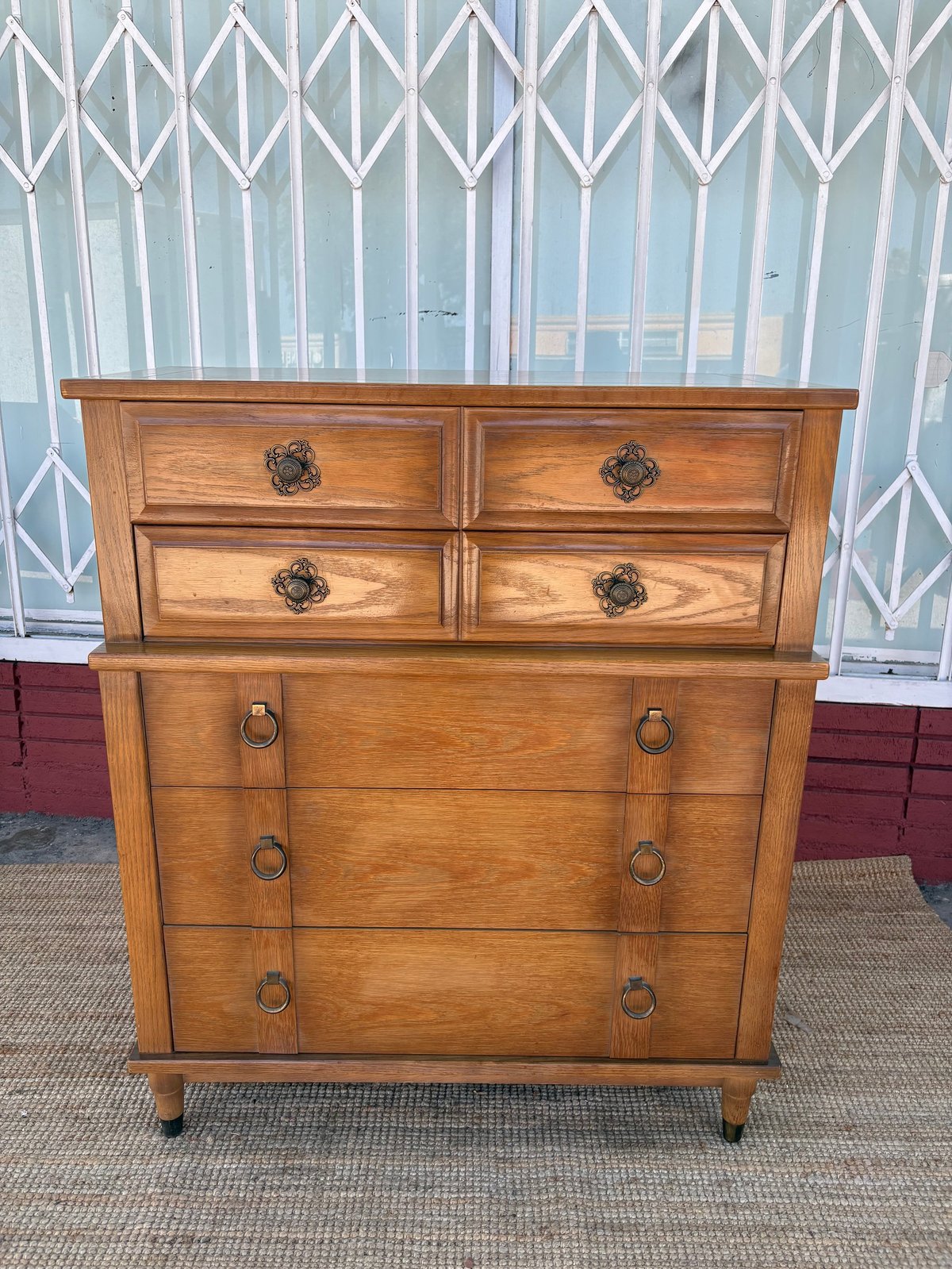 Vintage wooden chest of drawers with ornate handles, displayed outdoors against a white latticed fence with red brick visible nearby