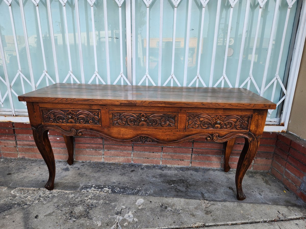 Ornate wooden console table with carved details and curved legs positioned against a window with sheer curtains