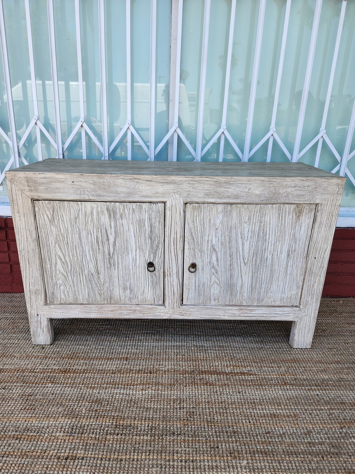 Whitewashed wooden console table with two cabinet doors and metal handles, placed on a textured area rug