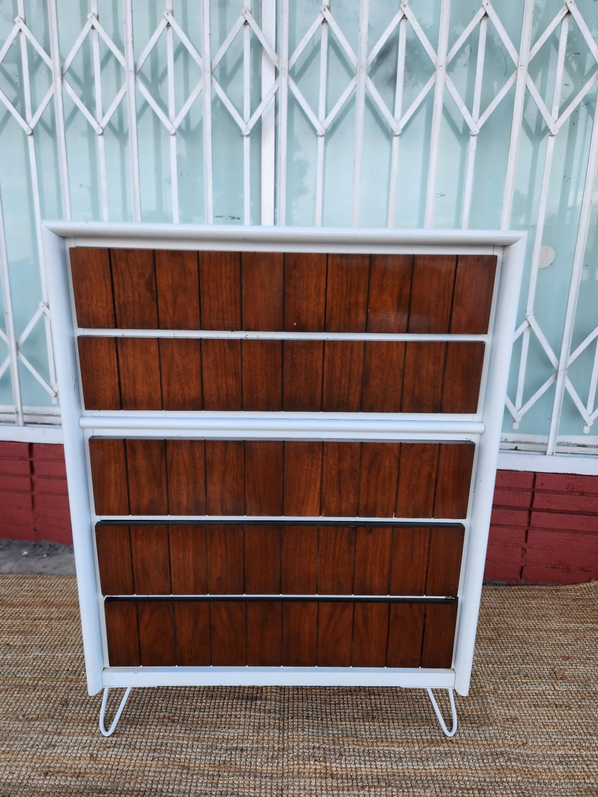 Mid-century white metal dresser with five brown wooden drawers and angled legs against a metal gate background