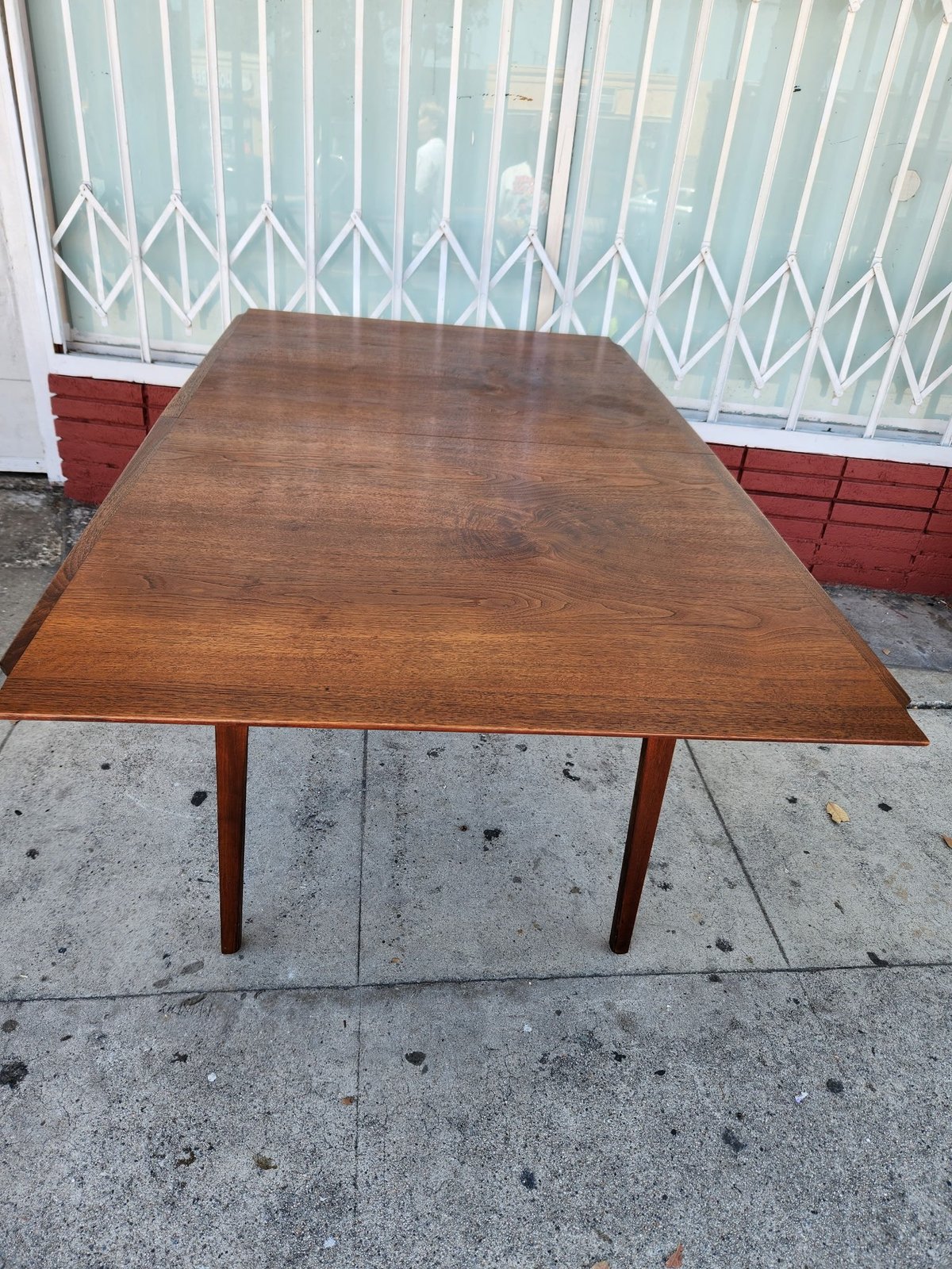 Mid-century wooden dining table with tapered legs on a sidewalk, white security gate visible in background