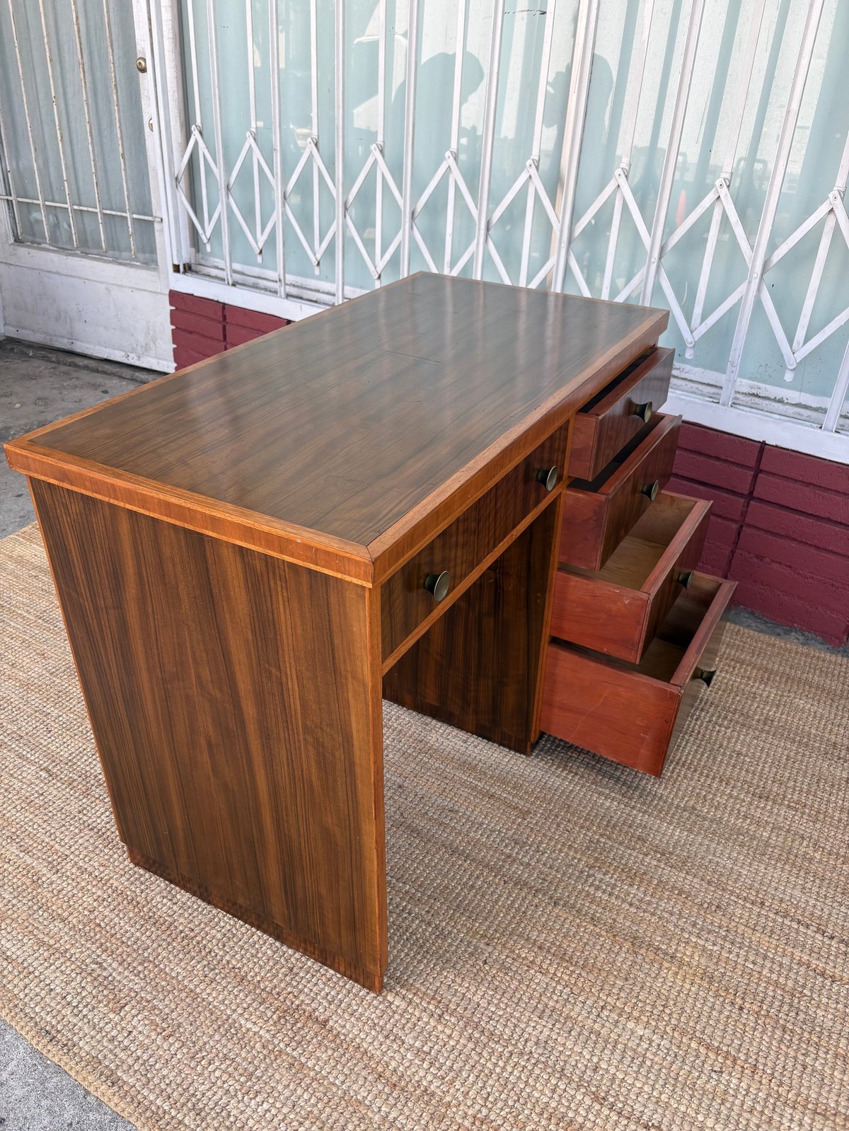Wooden desk with dark top and storage drawers on one side, positioned on a beige carpet with metal gate and red door in background