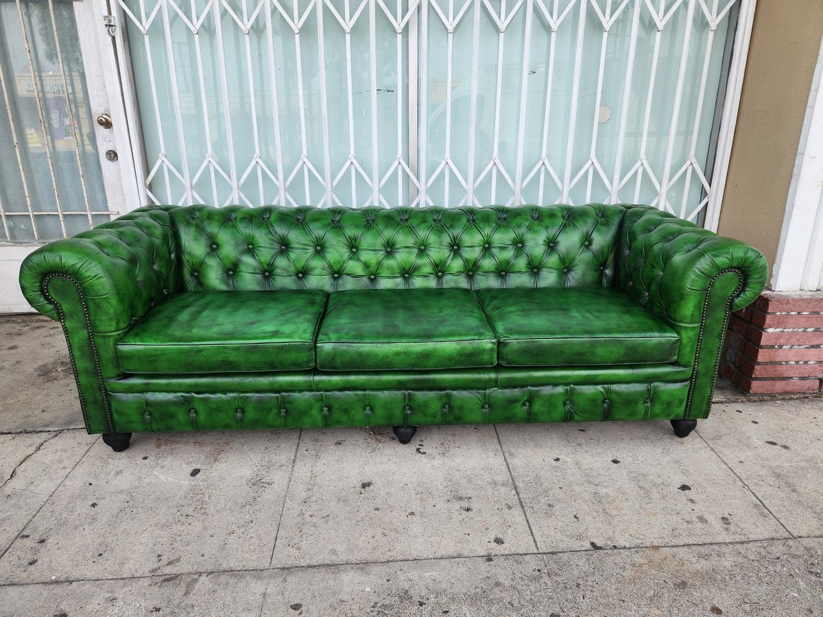 Green leather tufted Chesterfield sofa sitting on a concrete sidewalk in front of a white gate