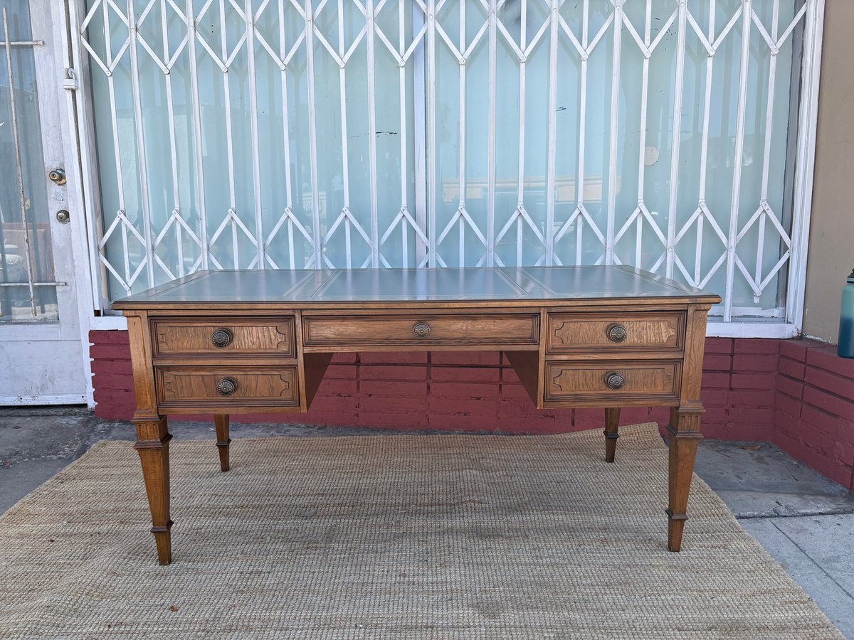 Vintage wooden desk with five drawers on tapered legs, positioned on a woven mat against a metal accordion gate