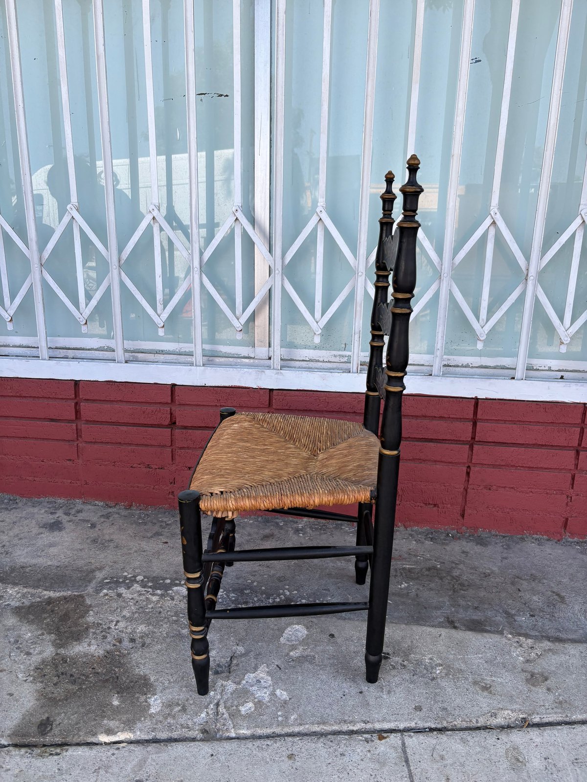 Black wooden chair with woven rush seat positioned on street pavement against a building with red and white shuttered windows