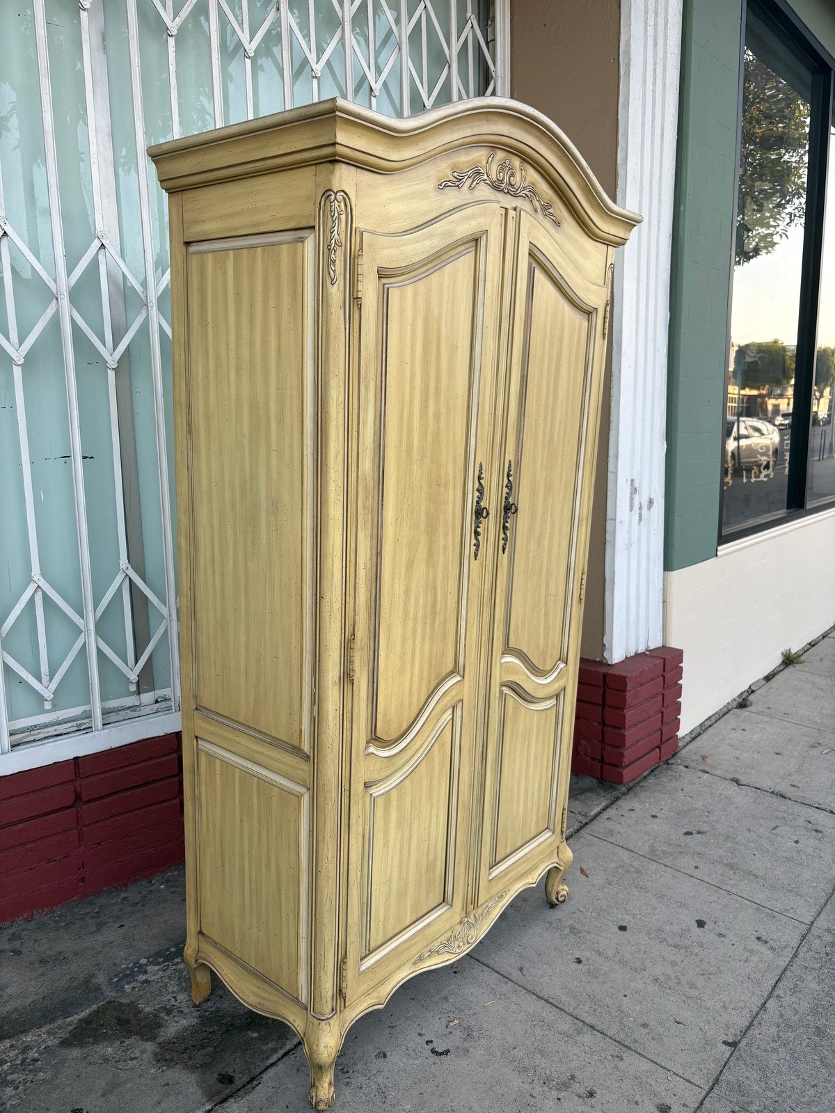 Antique cream-colored wooden armoire with curved top and ornate handles displayed on a sidewalk storefront