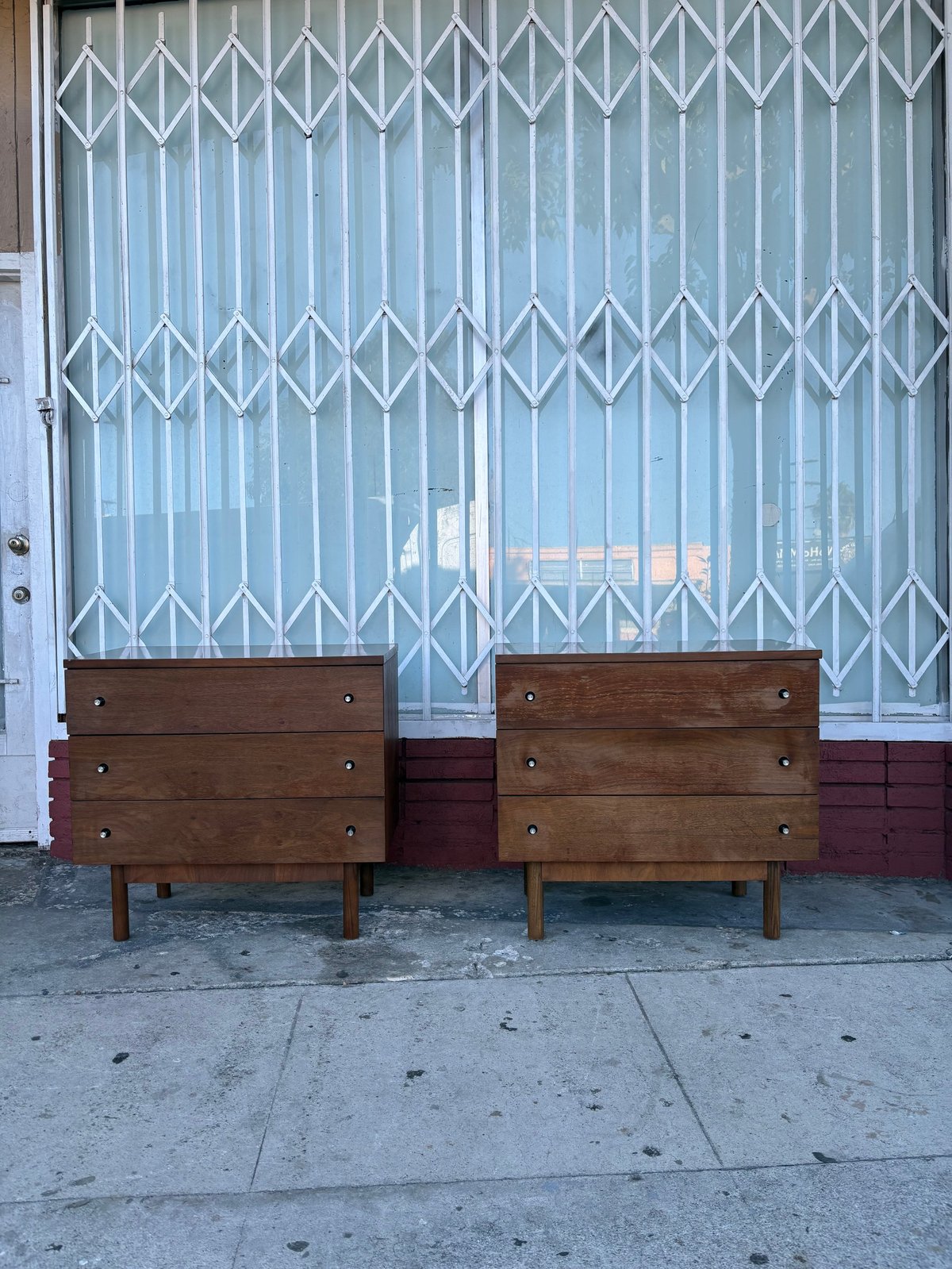 Two mid-century wooden dressers on a sidewalk in front of an accordion metal gate and storefront