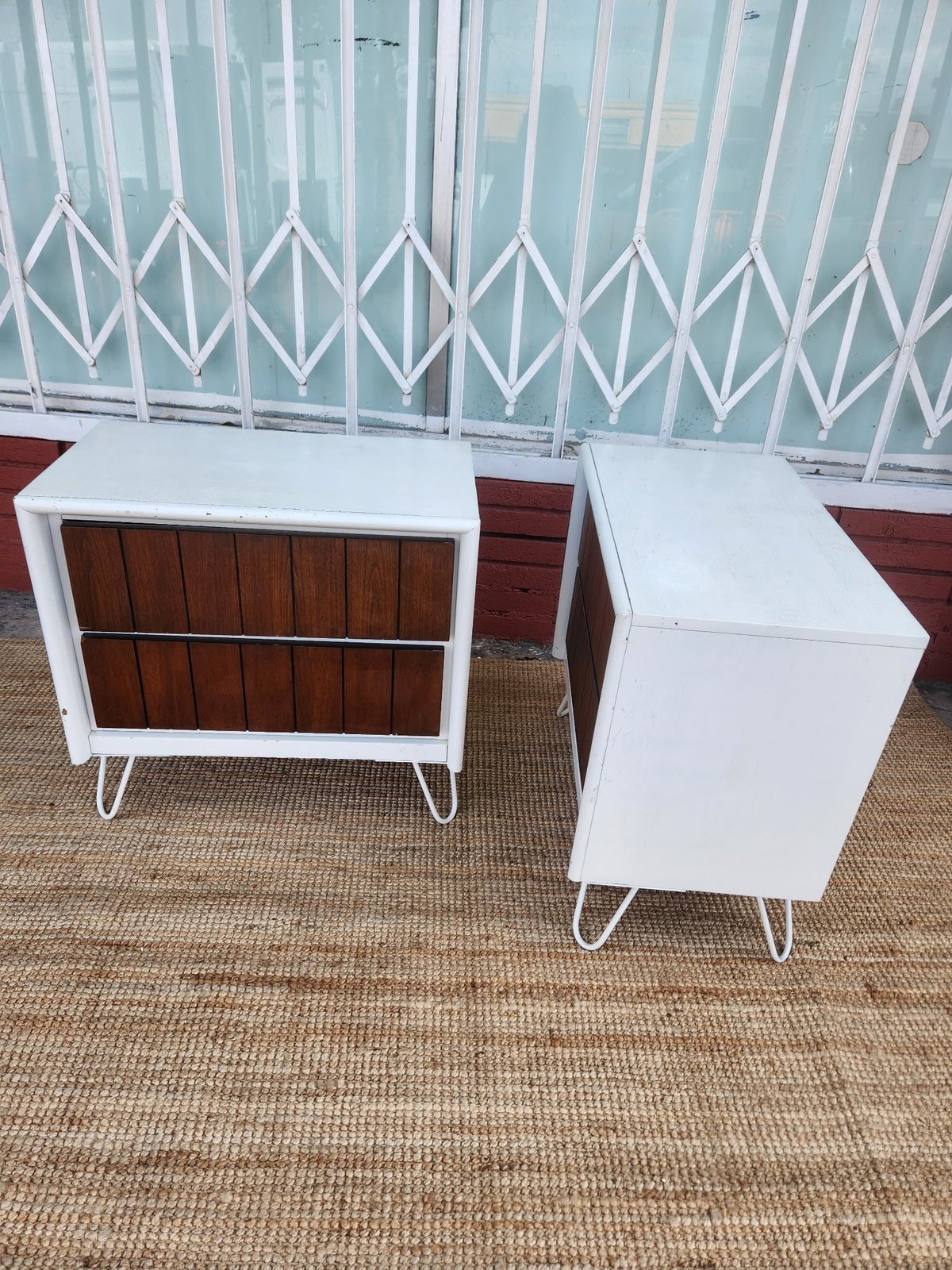 Two mid-century modern style nightstands with white frames and wooden fronts displayed on beige carpeting against a metal gate.